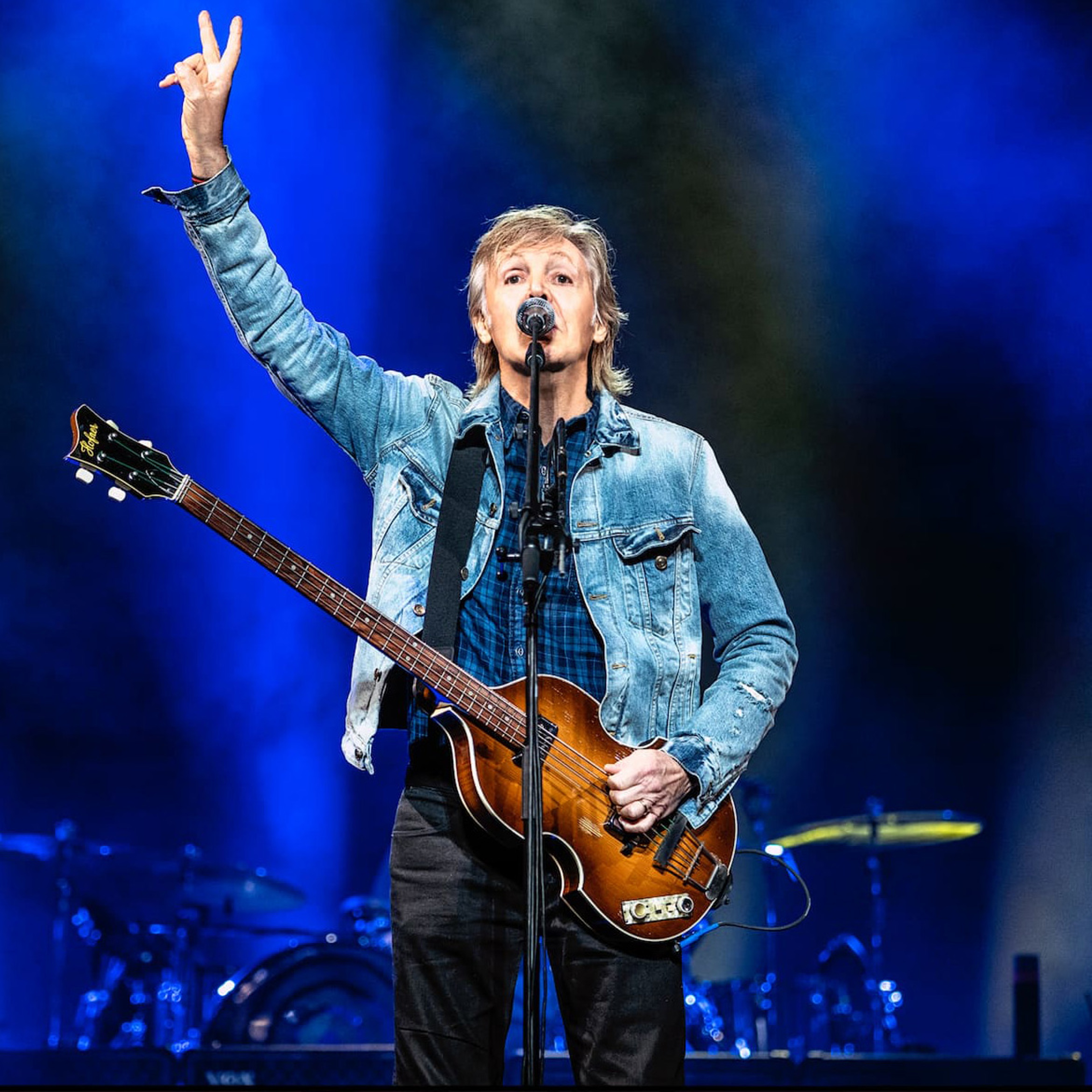 Photo of Paul performing in 2019 with his Hofner bass guitar and making a peace sign