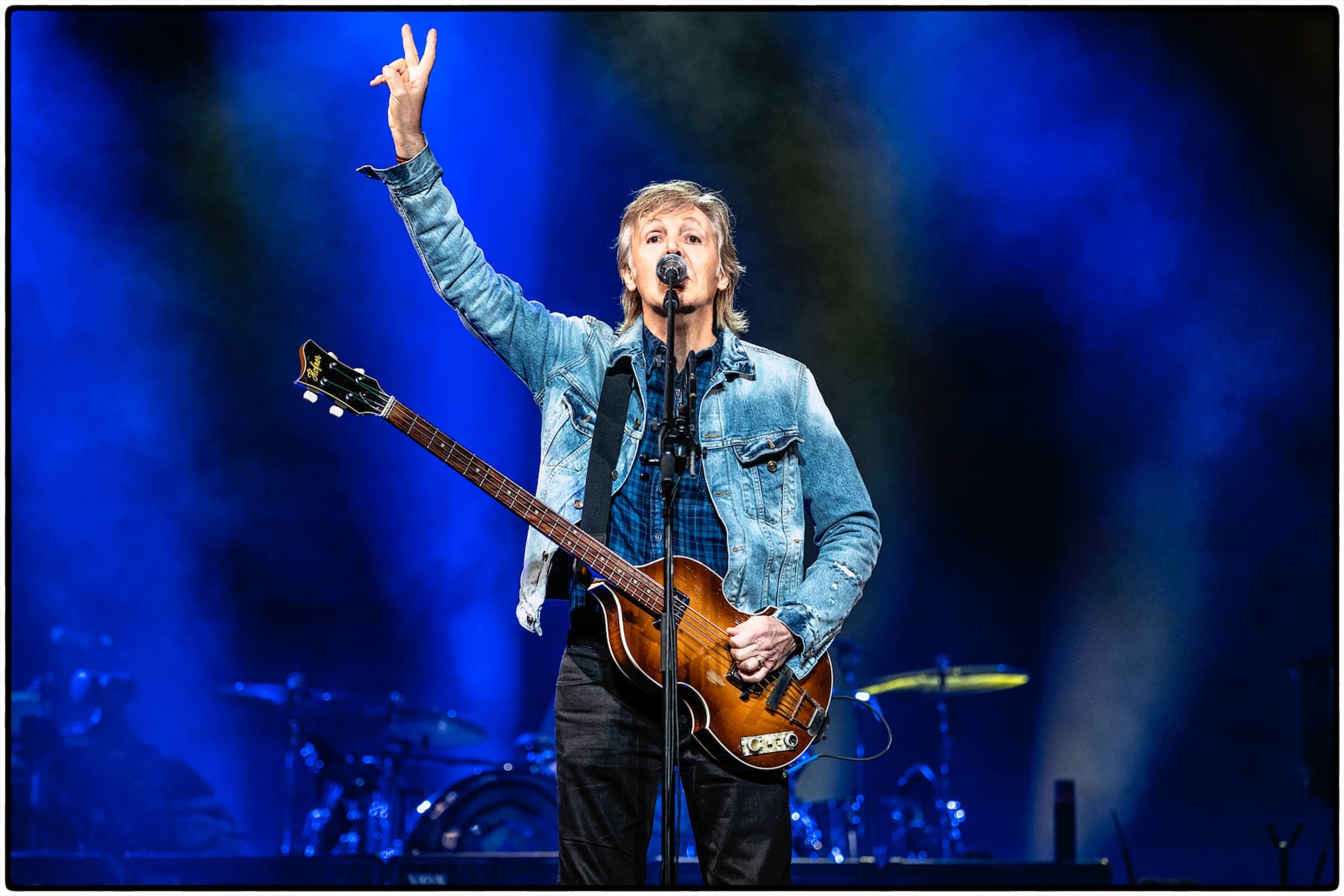 Photo of Paul performing in 2019 with his Hofner bass guitar and making a peace sign