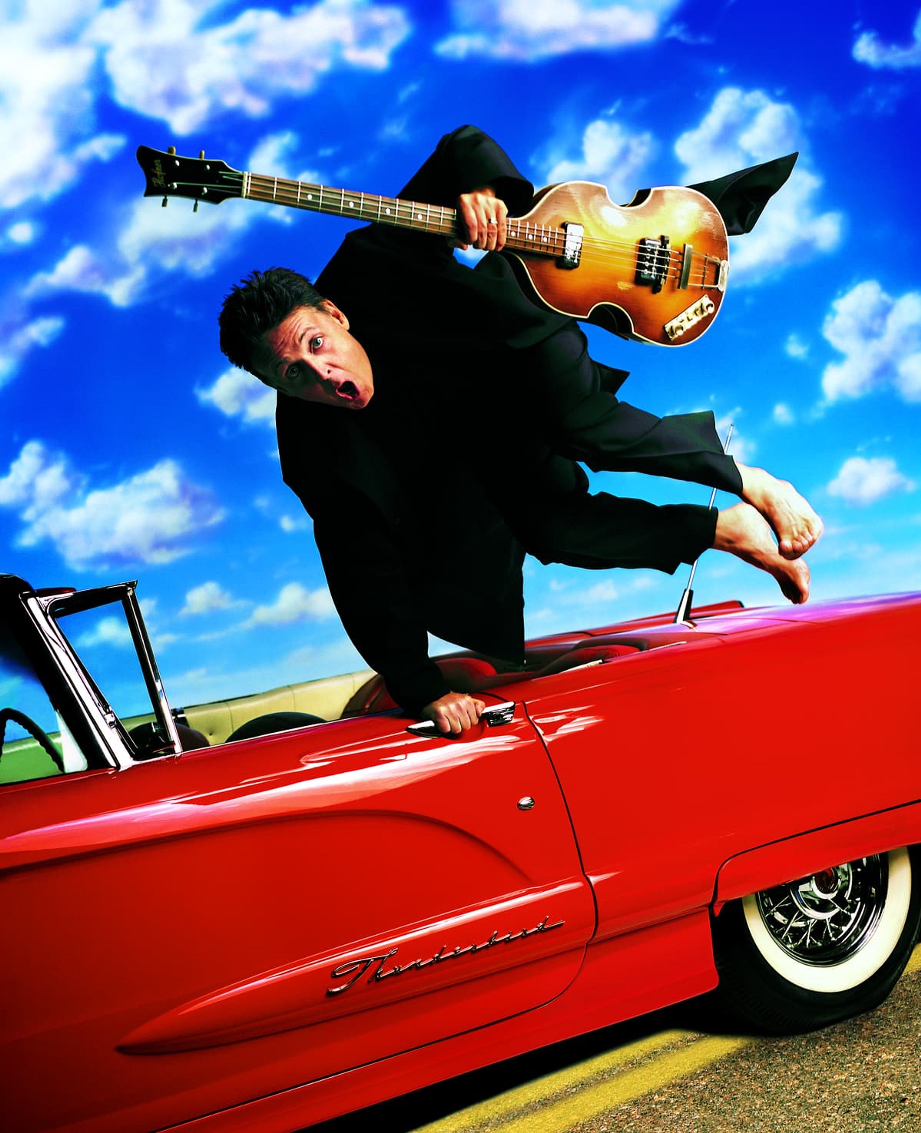 Paul jumping over a red car, holding a bass guitar