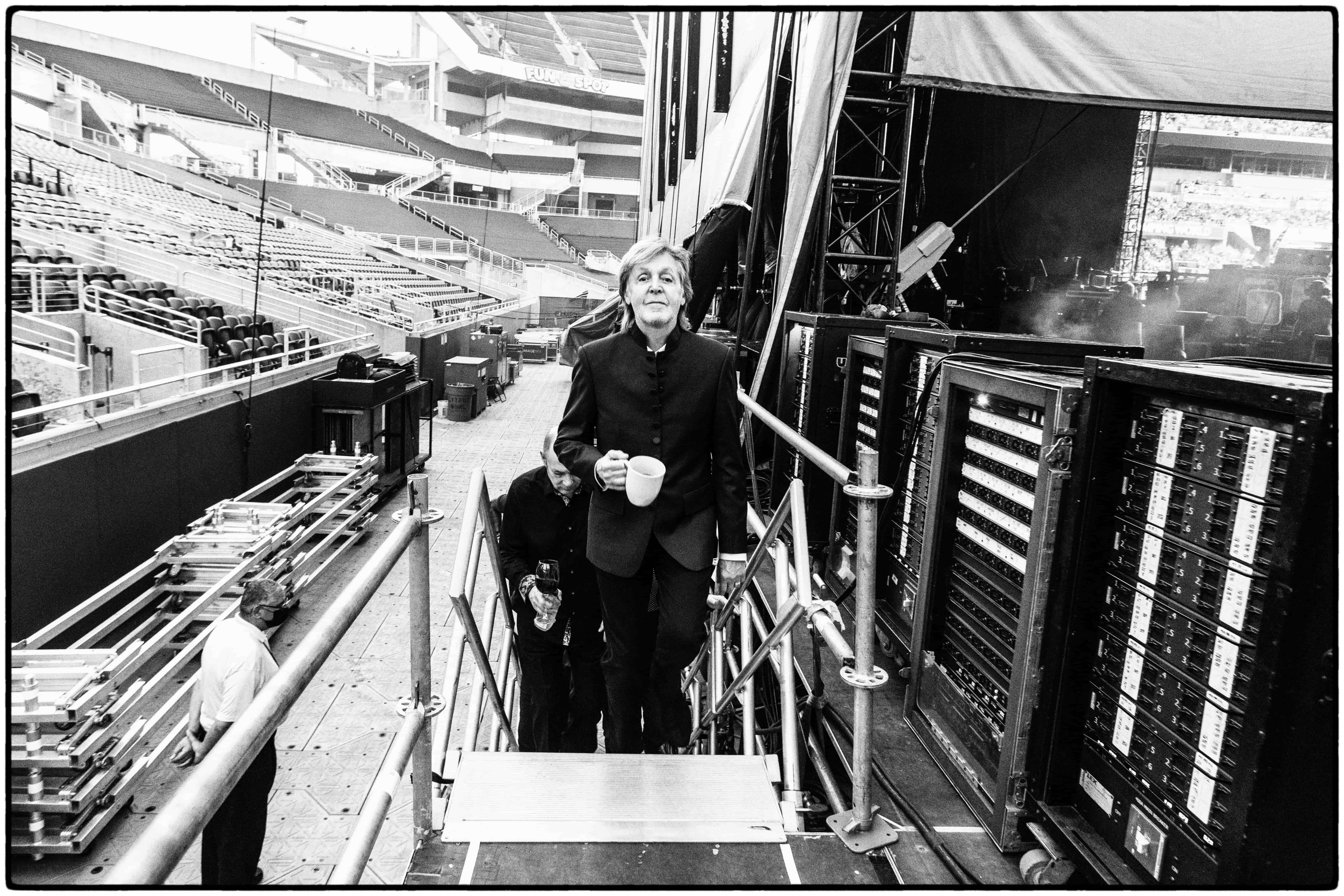 Black and white photo of Paul walking up steps backstage, holding a mug