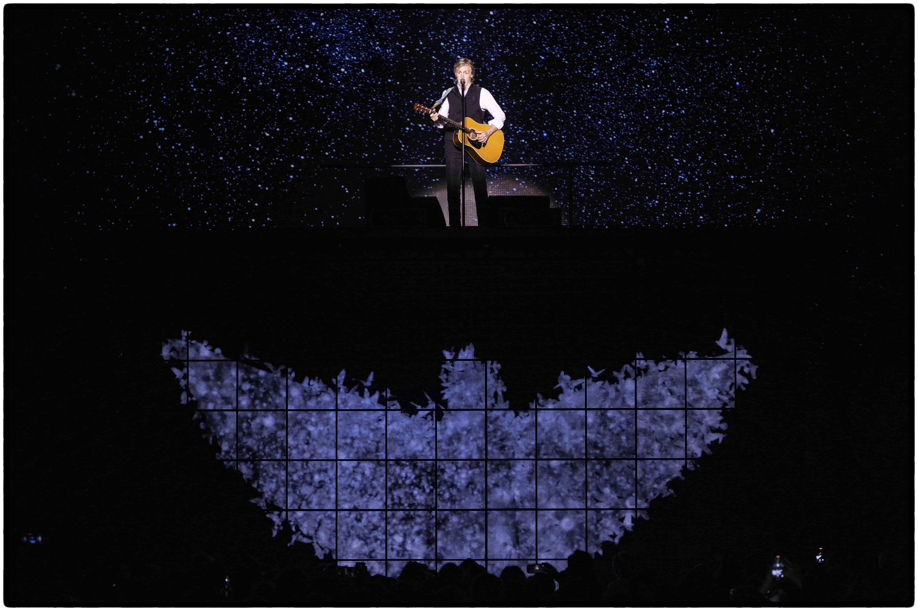 Paul playing guitar on a raised stage, with a light projection in the shape of a bird underneath him