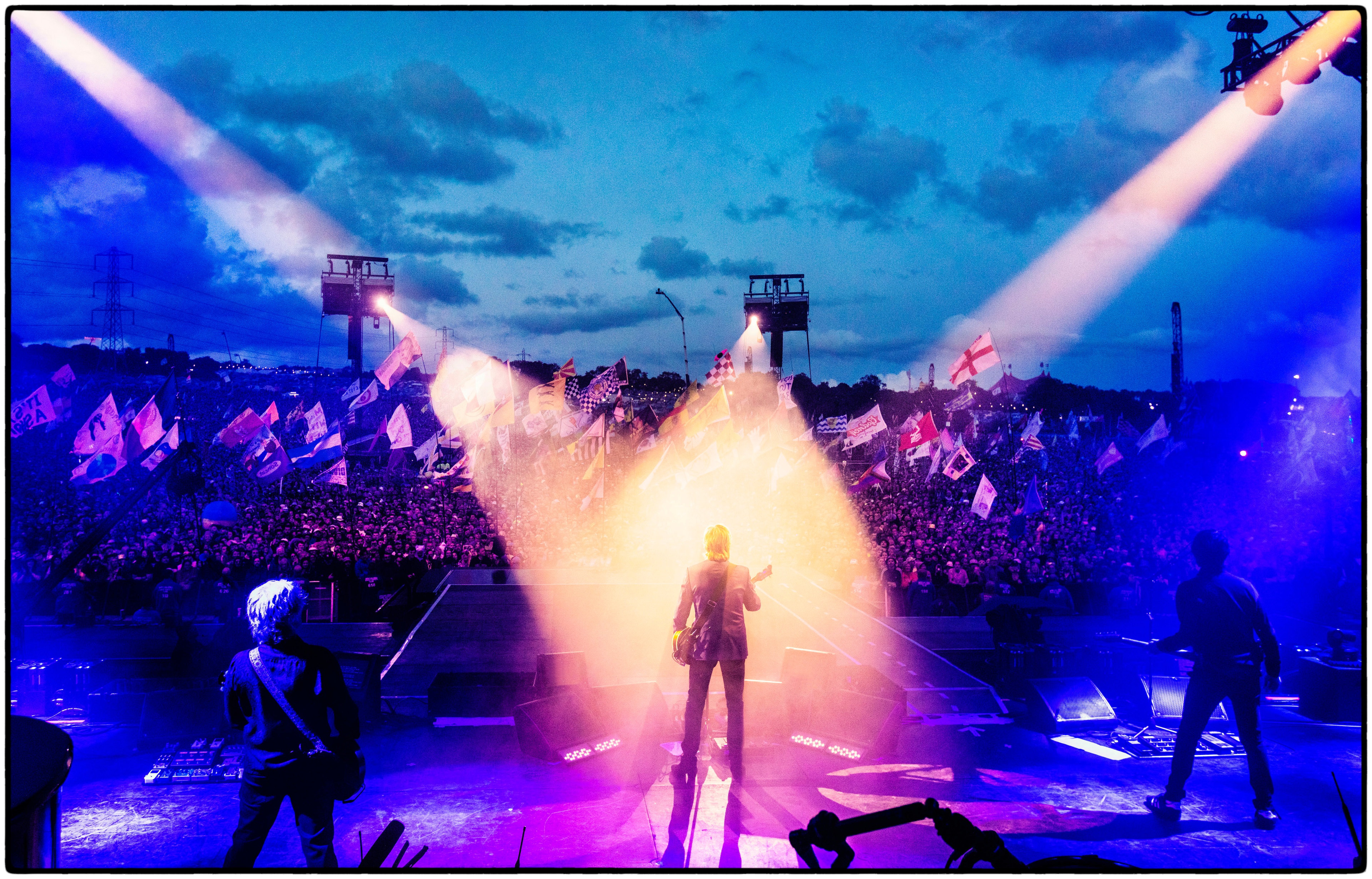Paul stands facing the crowd at Glastonbury, illuminated by a spotlight