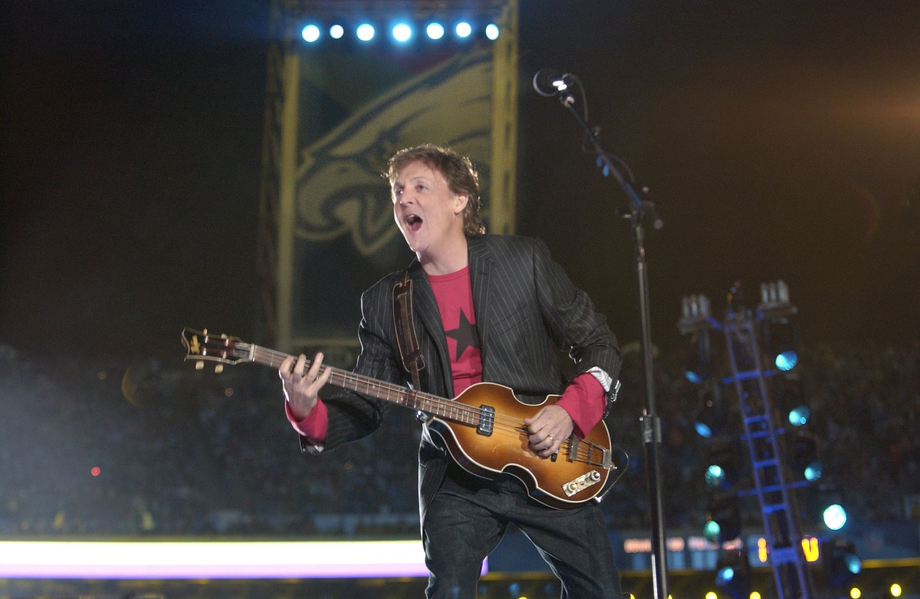 Paul wearing a red t shirt and black blazer, holding a bass guitar