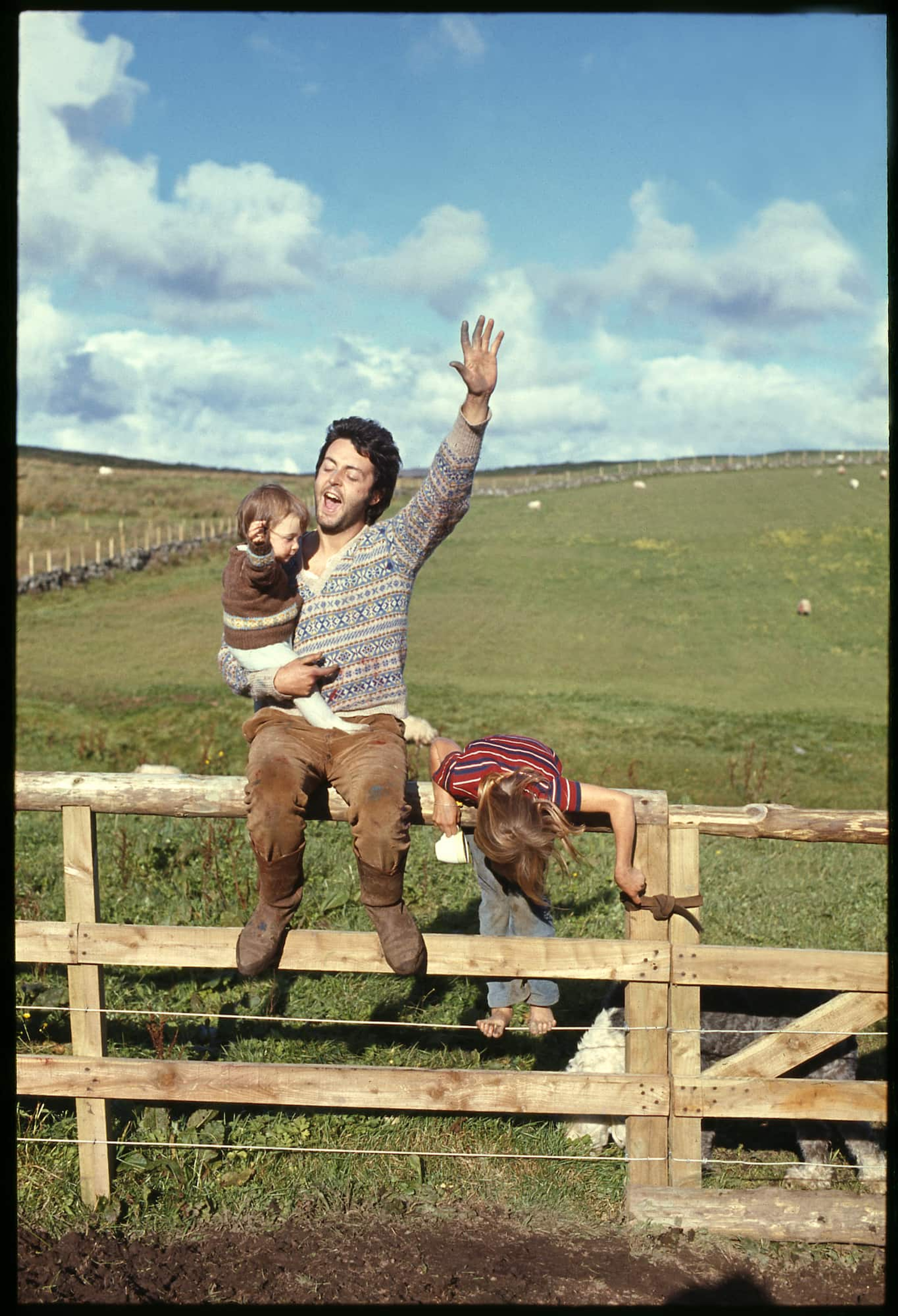 Colour portrait photo of Paul sat on a fence, holding Mary McCartney and with his arm in the air