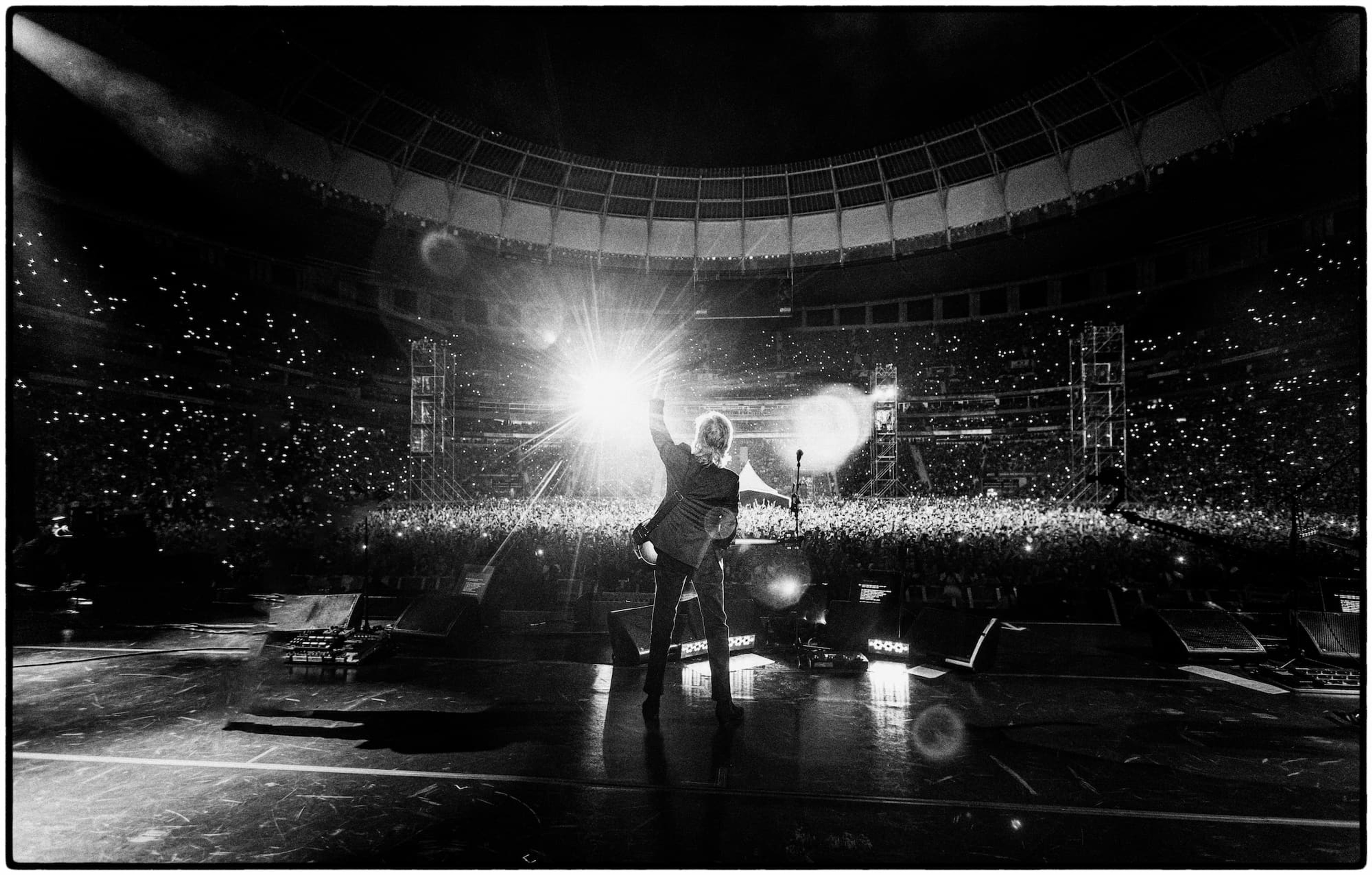 Black and white photograph of Paul McCartney raising his hand in the air while on stage in front of a sold-out crowd at Mané Garrincha Stadium in Brasília.