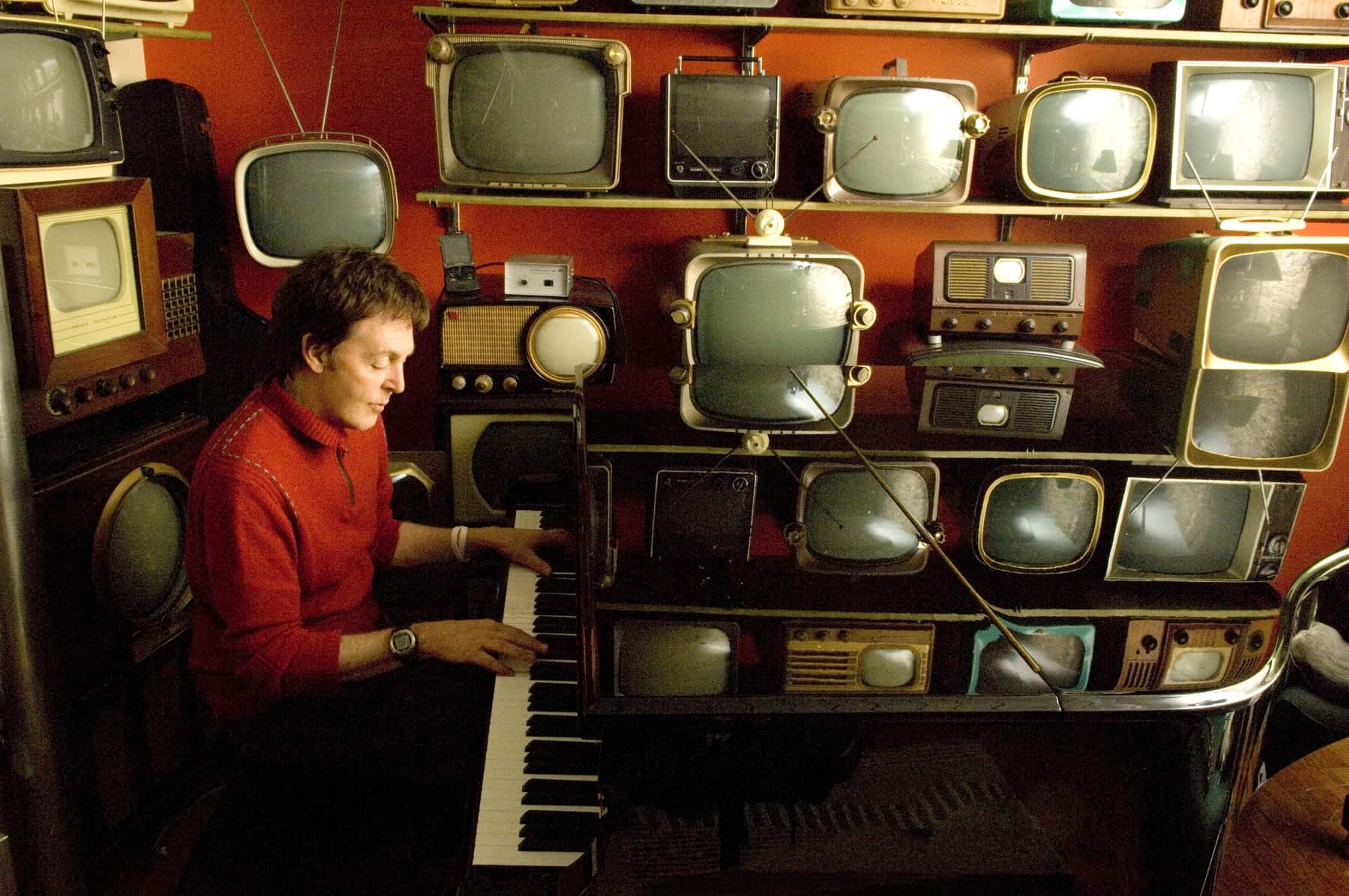 Paul sat at a piano with lots of old TVs on the wall behind him