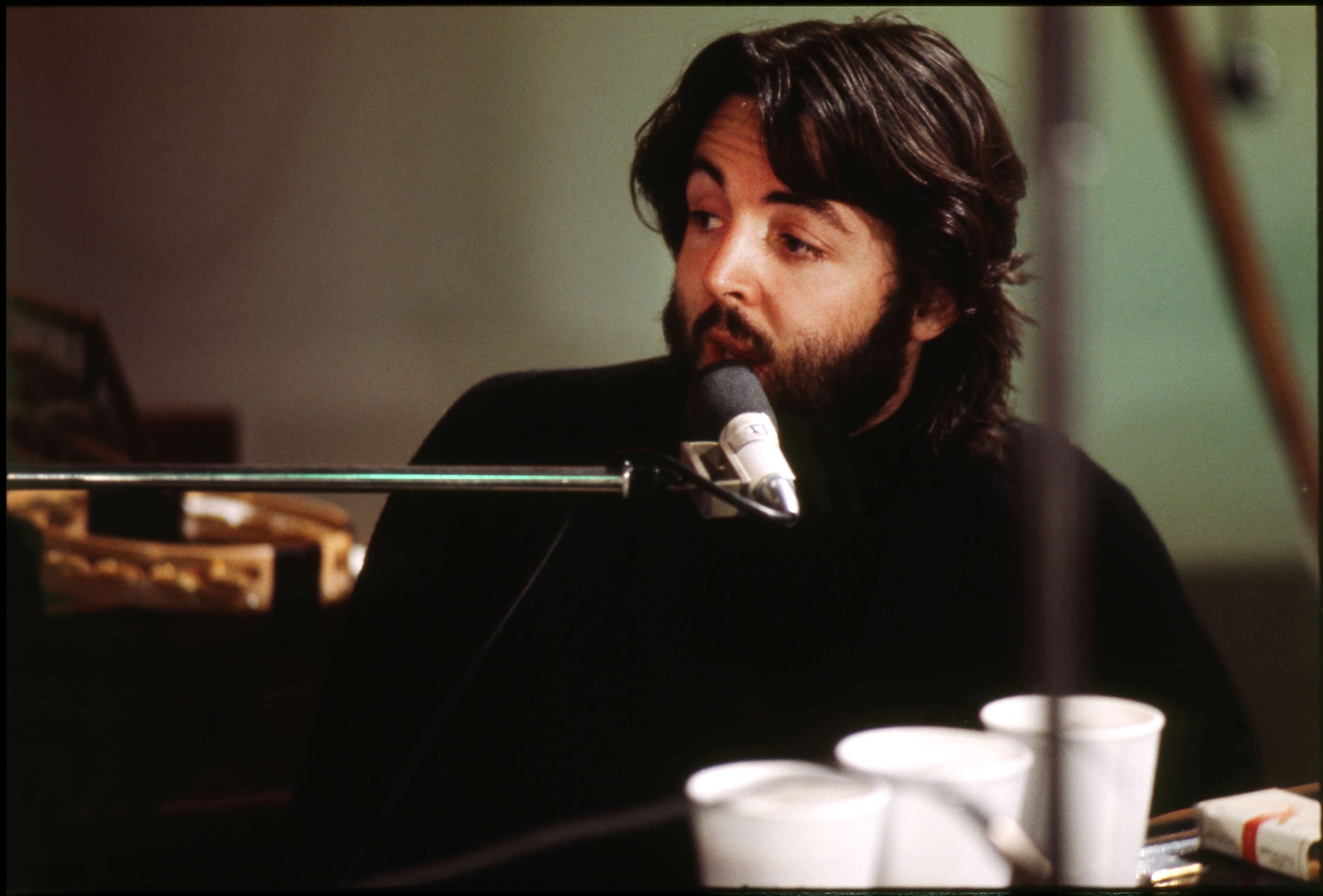 A bearded Paul sits at a piano singing into a microphone, with cups of tea balanced in front of him