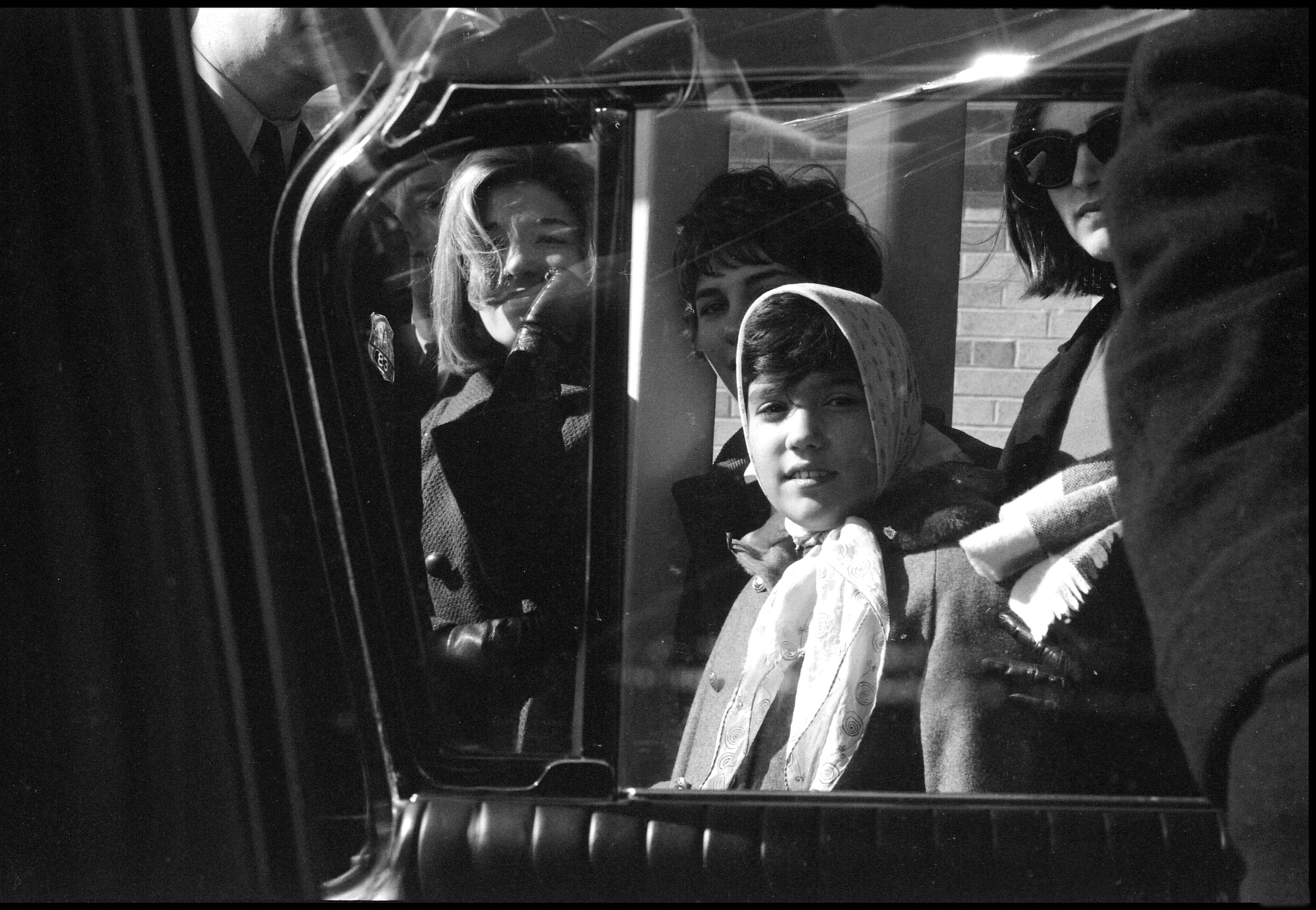 Black and white photo of a young girl wearing a headscarf, looking into a car window