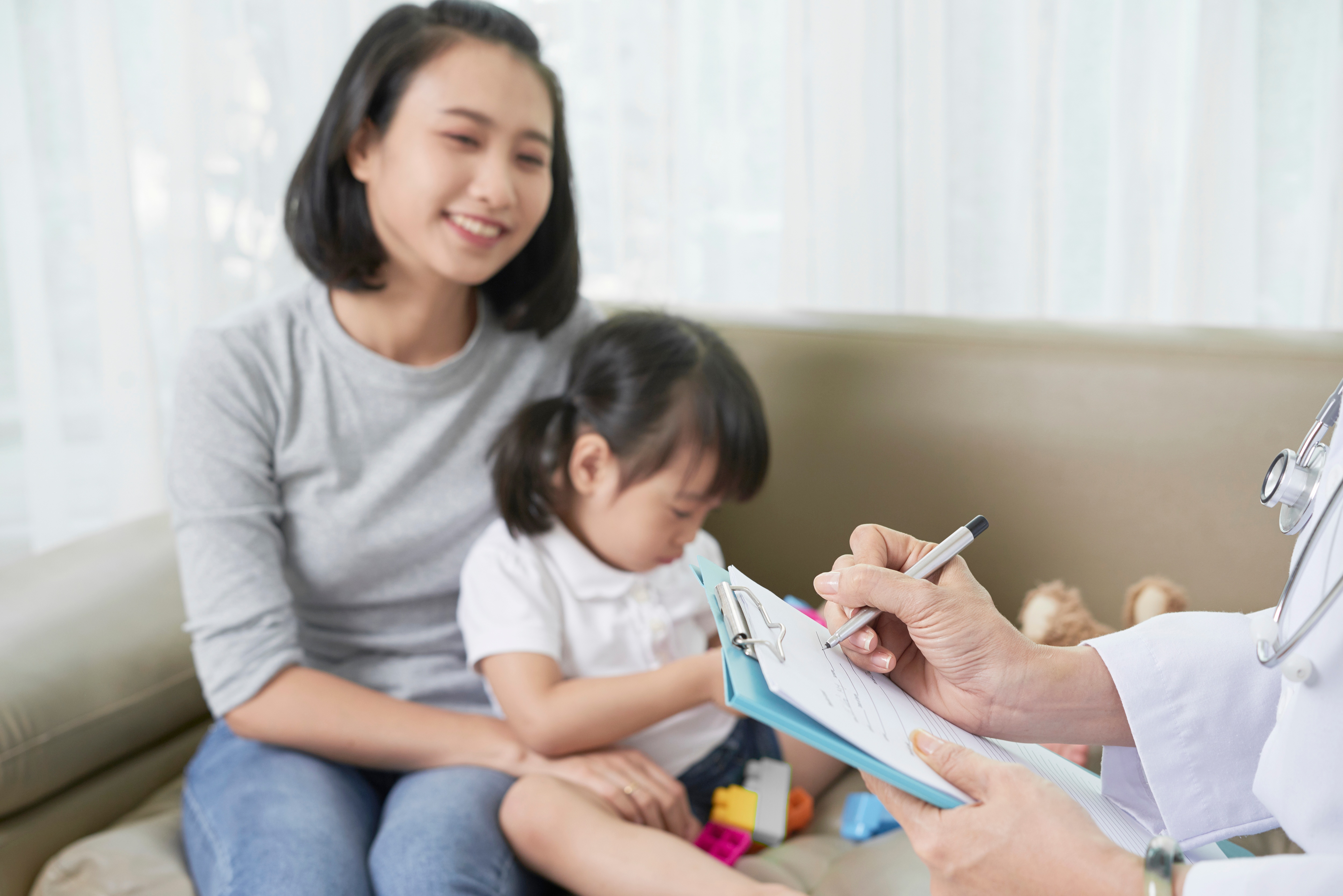 Young woman holds her toddler daughter on a sofa affectionately, with a medical professional filling up a form in the foreground