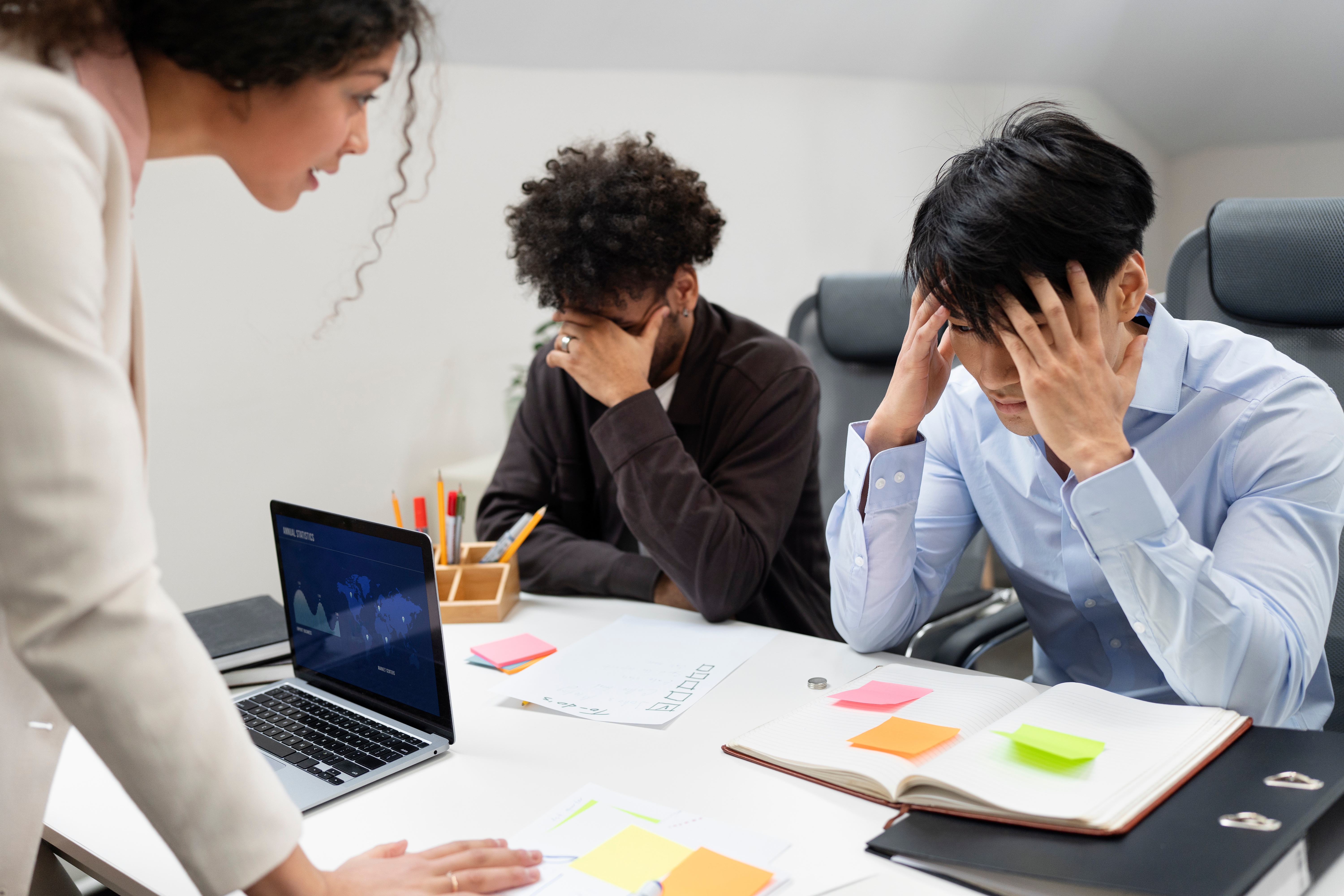 A woman sternly addresses two distressed male colleagues at a desk, indicating a tense and high-stress situation