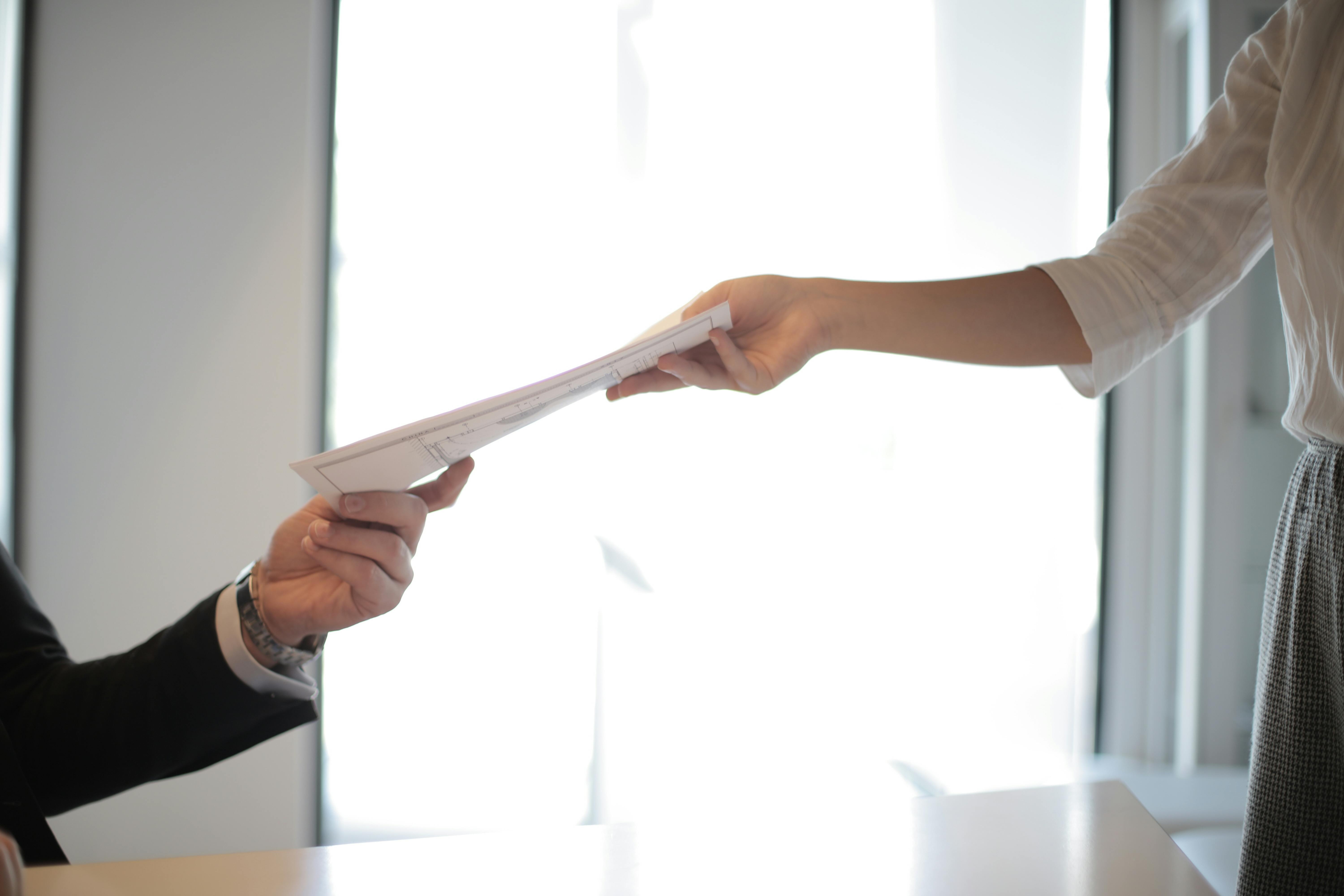 A woman in a white blouse hands a document to a man in a black suit.