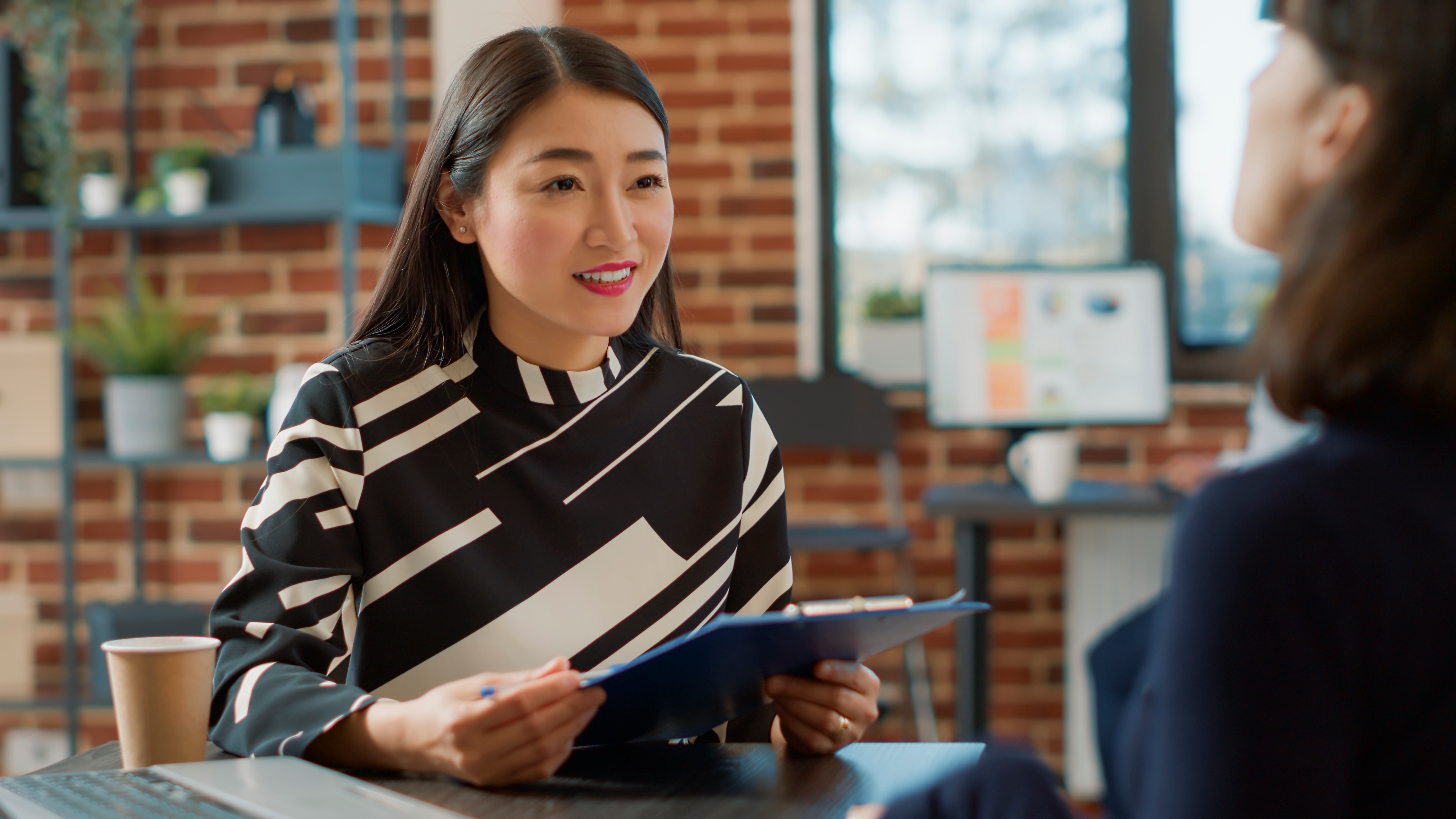 A female professional smiles while reassuringly at another woman opposite her, holding a clipboard in a modern office setting