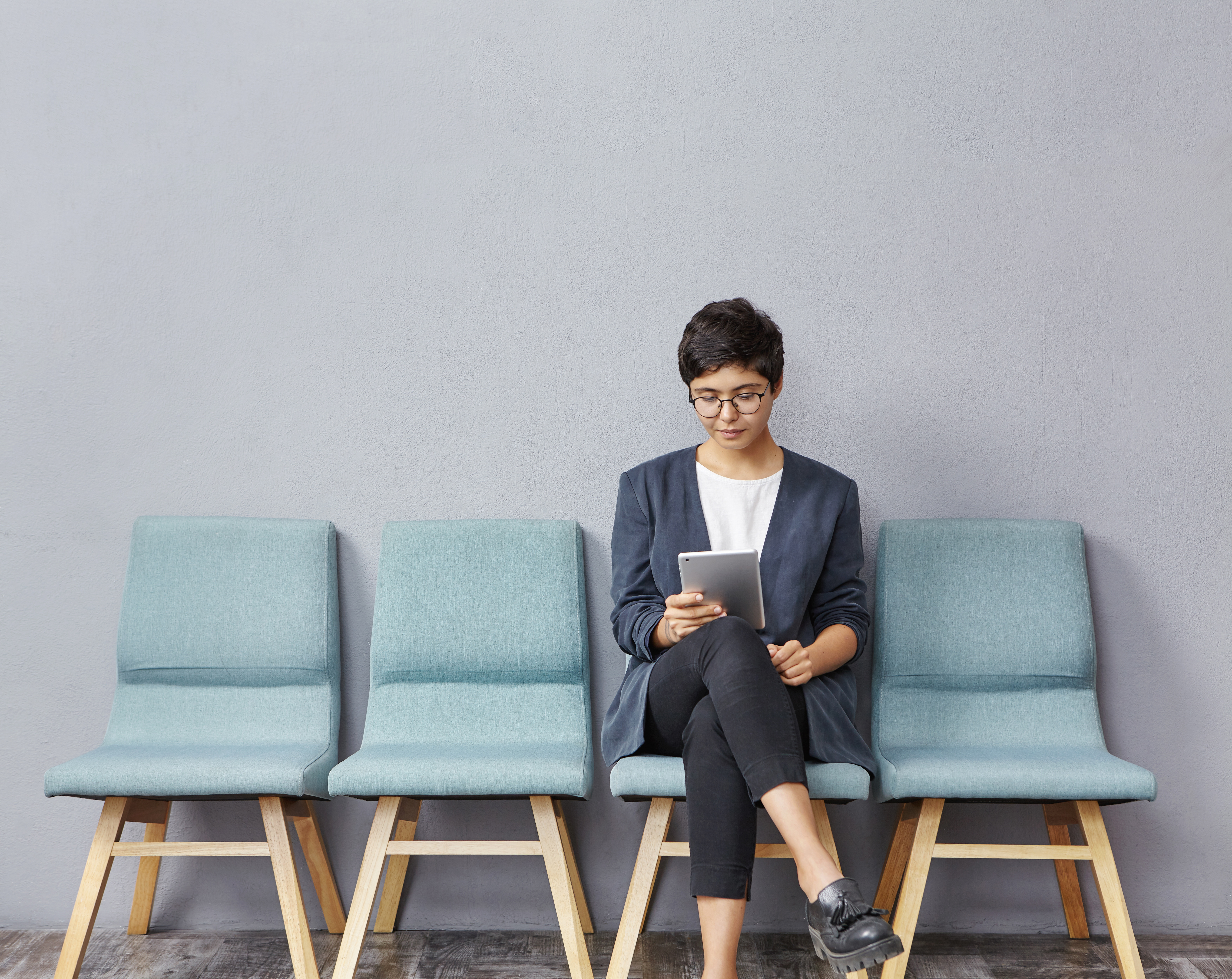 A young woman sits in a waiting area, reviewing a tablet, preparing to address selection criteria before her job interview.