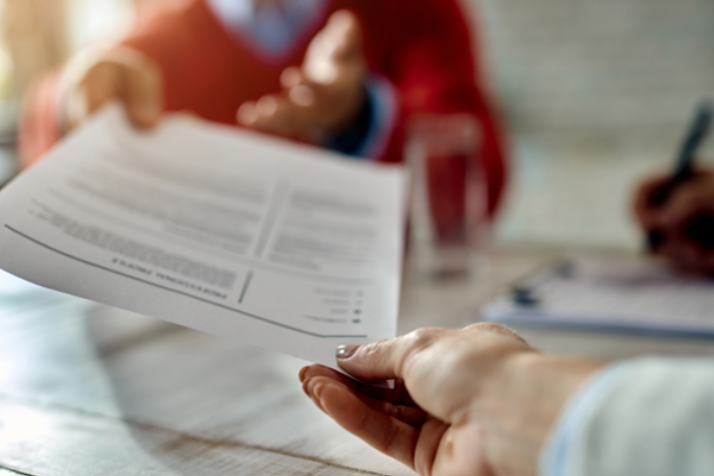 Close-up of a job applicant handing over a document, likely a resume or personal reference, during an interview in a work setting.