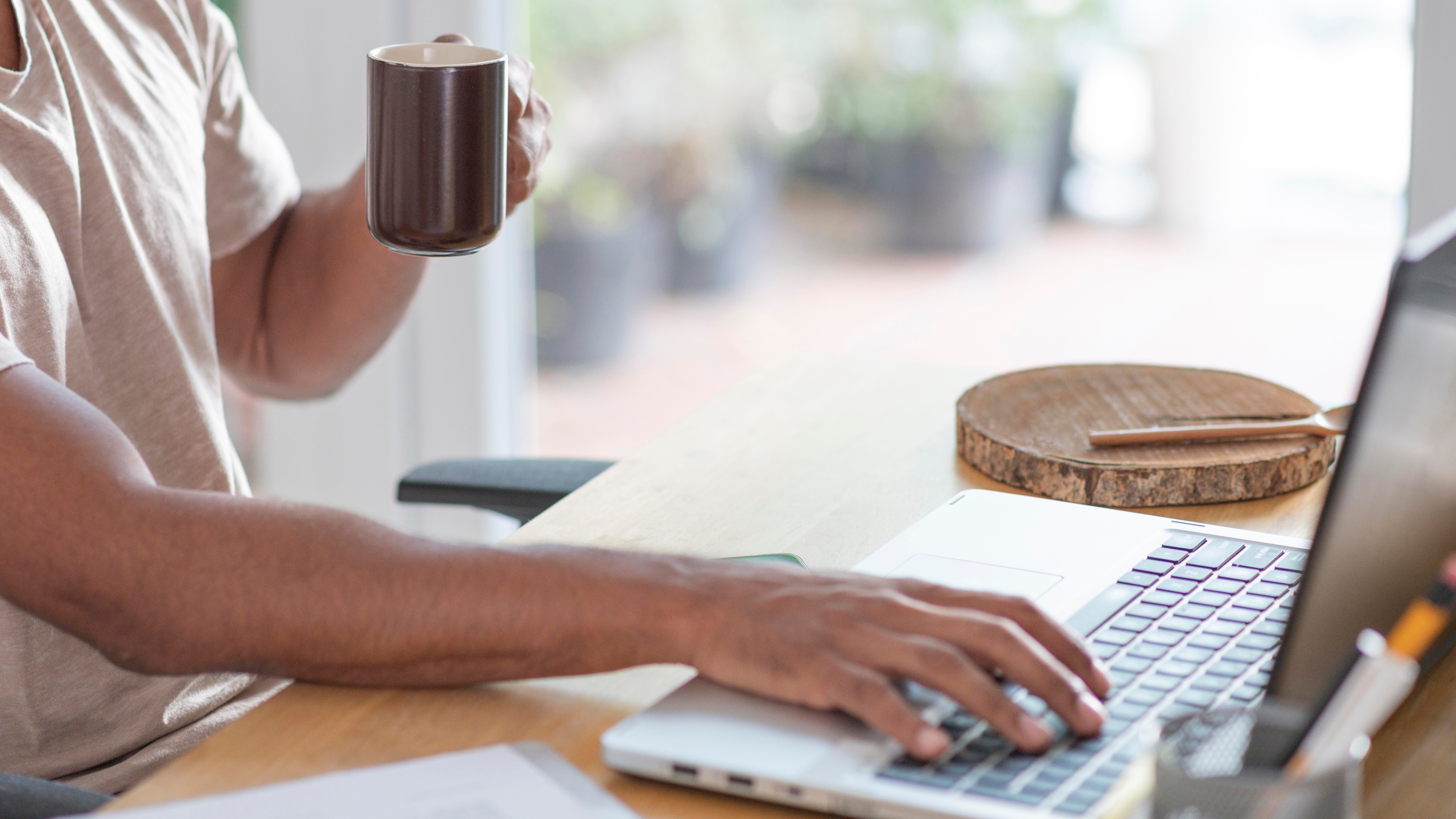 Man typing on laptop with right hand while holding a cup in left hand