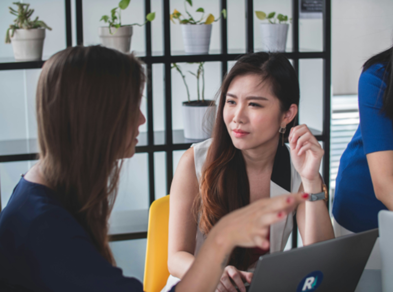 Two executives engage in a serious discussion at a desk, possibly about providing a personal reference, with a laptop and potted plants in the background.