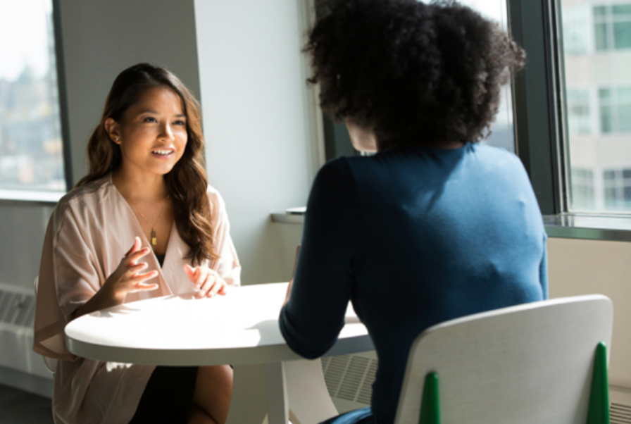 Two women sit across from each other at a table in a bright office, engaged in a discussion, possibly about providing a personal reference for a job application