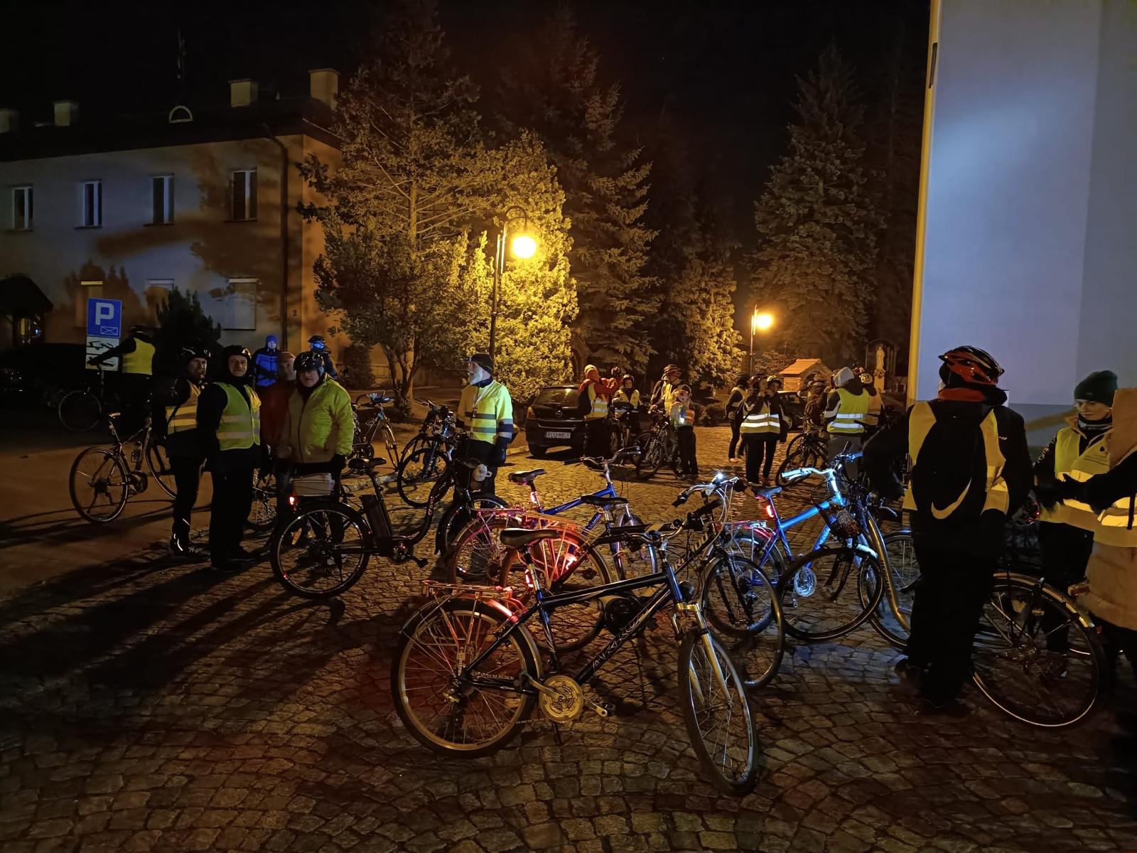 Rowerowa Droga Krzyżowa | Rowery zaparkowane na brukowanym placu, między nimi kilku kolarzy. W tle palące się latarnie, drzewa, budynek plebanii.jpg