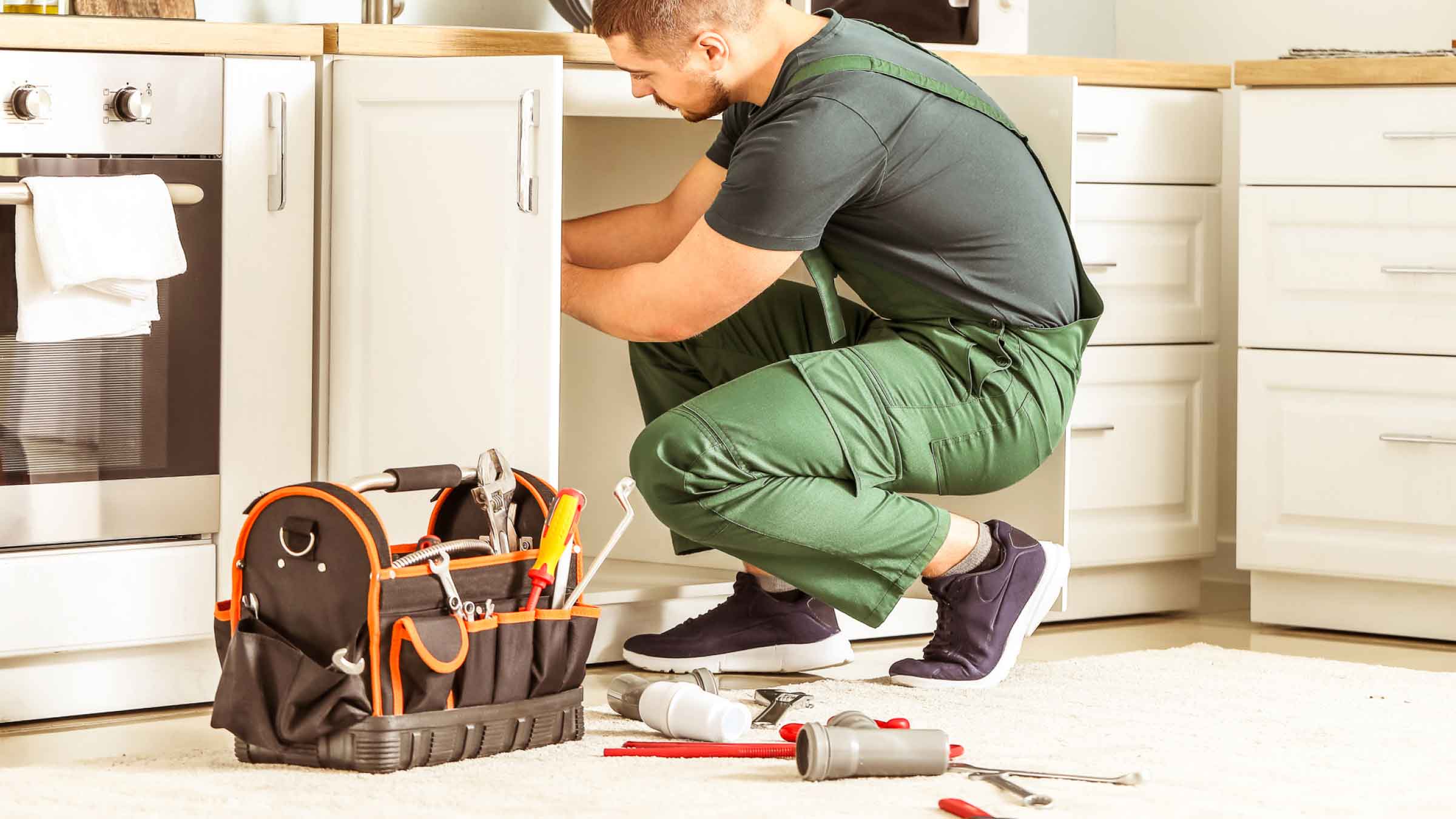Plumber in green overalls crouches in front of kitchen sink cupboard, his tools and pipe parts are laid out beside him.