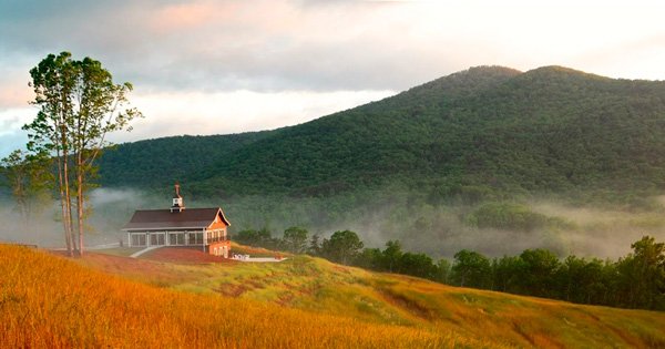A rustic cottage sitting on a hilltop in northern Georgia. There is a green mountain in the background and the cottage is surrounded by fog. The time of day looks to be the early morning.