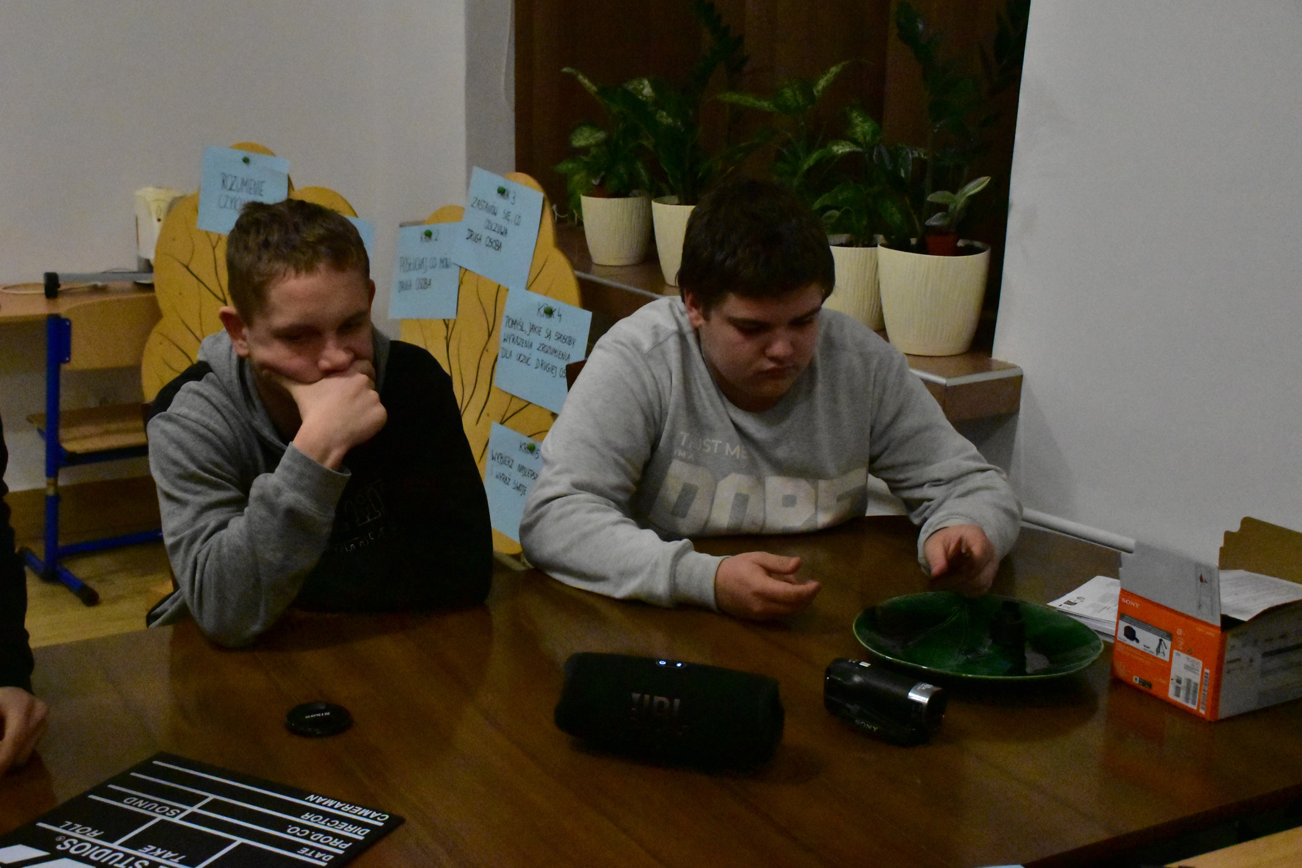 Cykl zajęć z programu „Fajna ferajna”  | Dwaj chłopcy siedzą za stołem, jeden podpiera dłonią twarz. Przed nimi na blacie głośnik, kamerka oraz klaps filmowy. Obok ceramiczna miska i pudełko.JPG