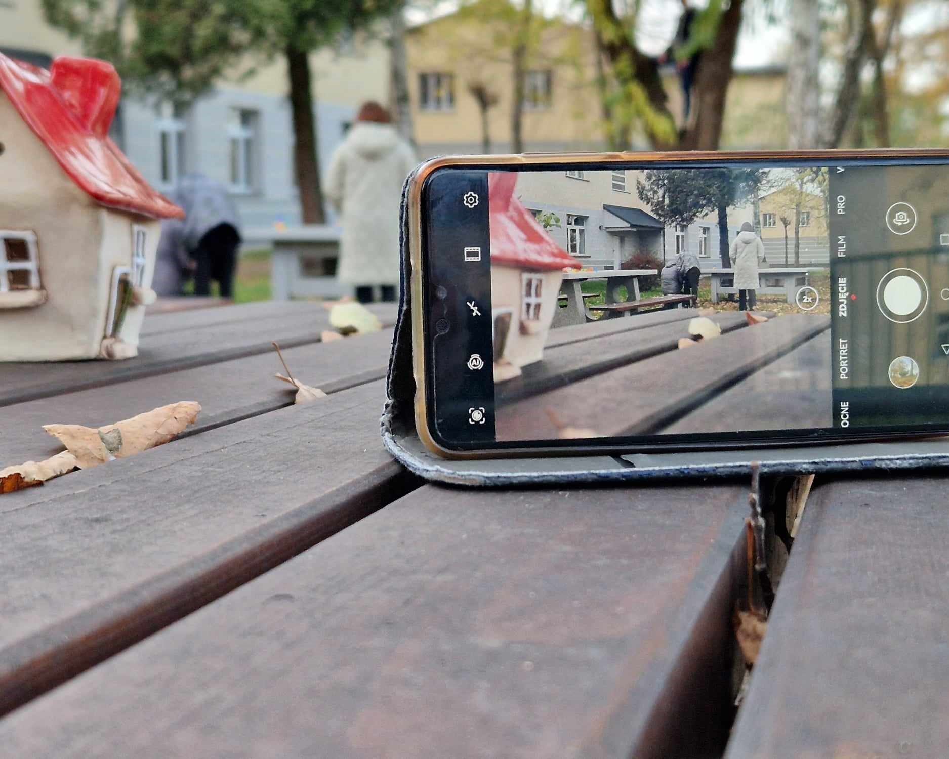 Cykl zajęć z programu „Fajna ferajna”  | Telefon komórkowy z włączonym aparatem stoi na deskach, na ekranie widać ceramiczny domek, sylwetki ludzi, drzewa, budynki. Jest to jakby zdjęcie w zdjęciu.jpg