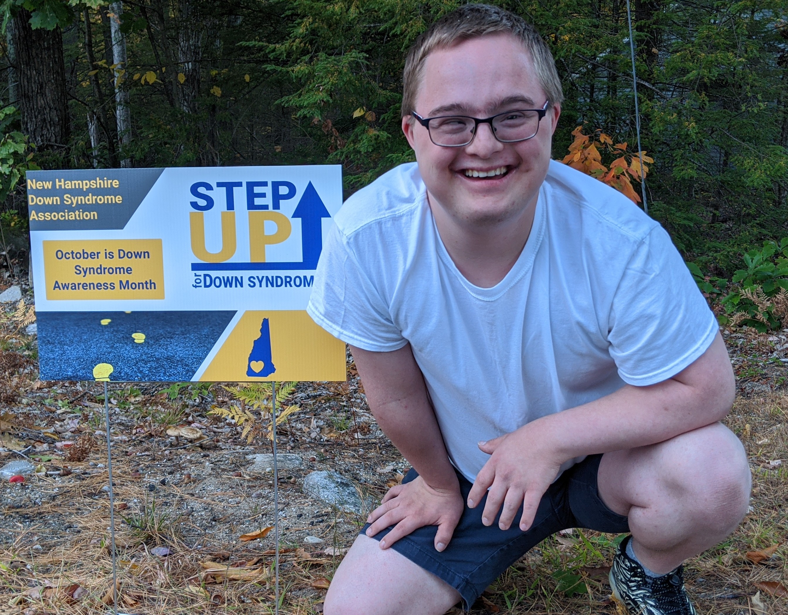 A young man with Down syndrome smiling in front of a sign promoting Down syndrome awareness month in October