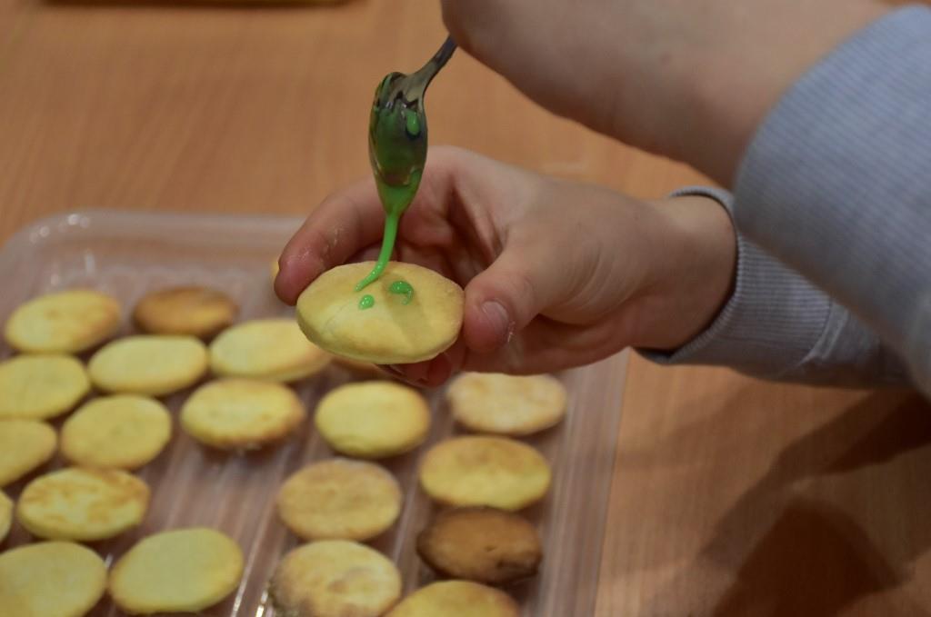 Dzień Św. Patryka  | Zbliżenie dłoni z ciasteczkem i łyżeczką u góry. Z łyżeczki na ciasko ścieka zielony lukier. W tle taca z resztą ciastek.JPG