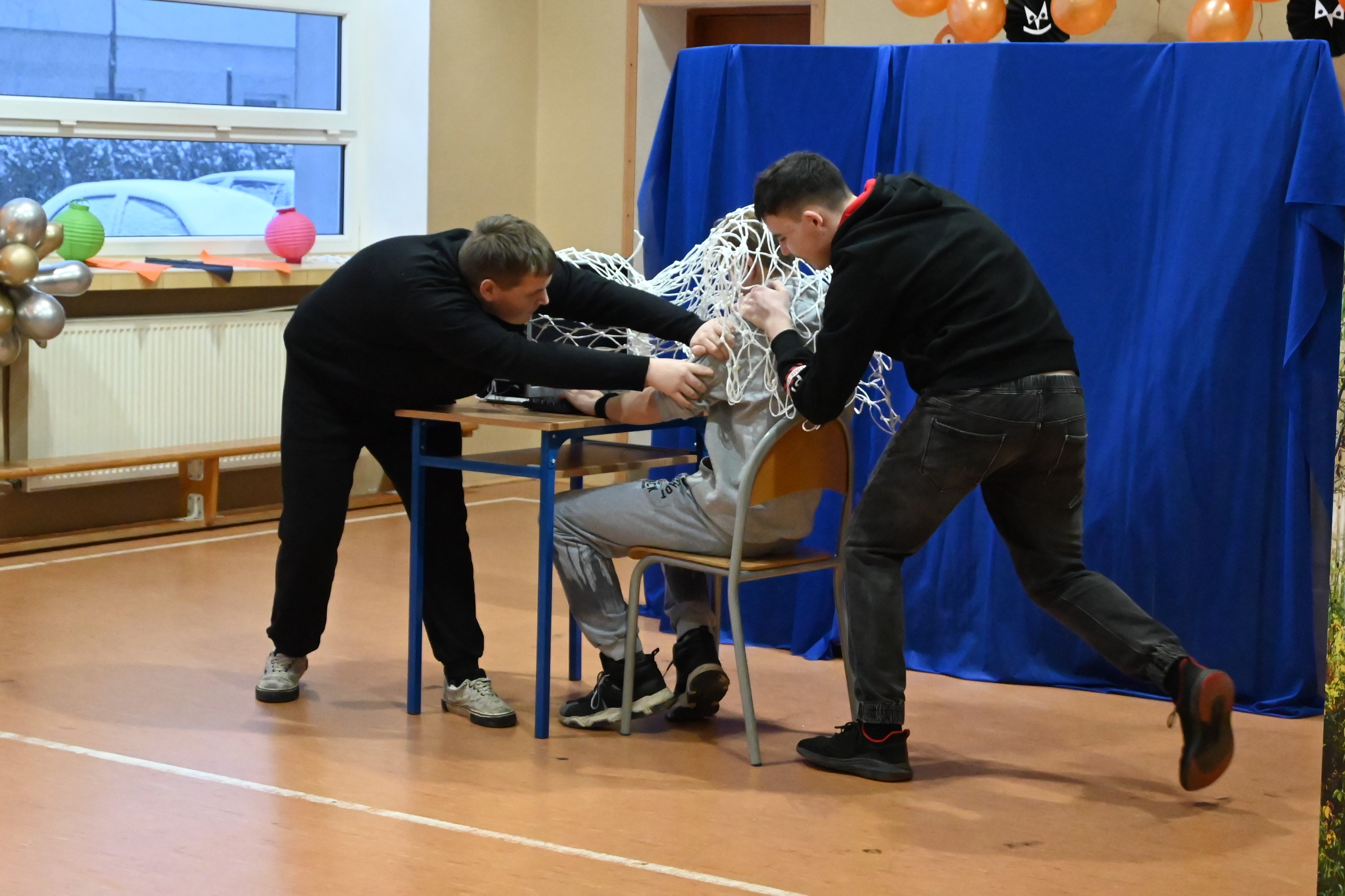 O uzależnieniach językiem teatru | Chłopiec siedzi przy stoliku, na nim narzucona biała sieć. Dwóch innych chłopców, jeden z przodu, drugi z tyłu, próbują przeciągnąć siedzącego k.JPG