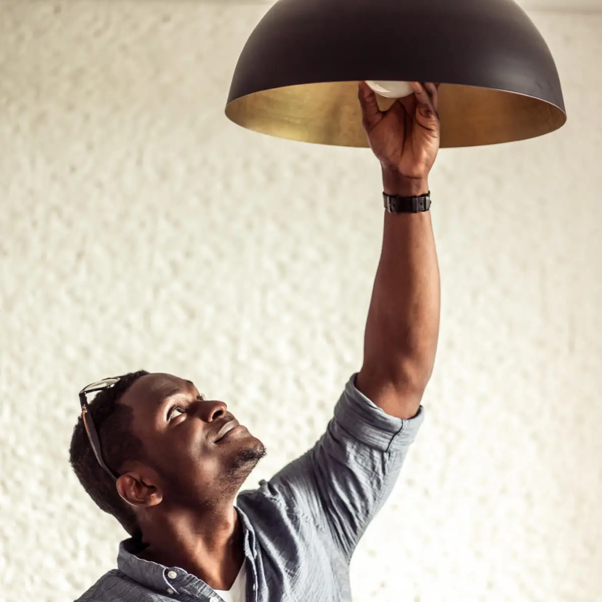 Young smiling man with glasses perched on top of his head, reaches up to a ceiling light to replace it's bulb.