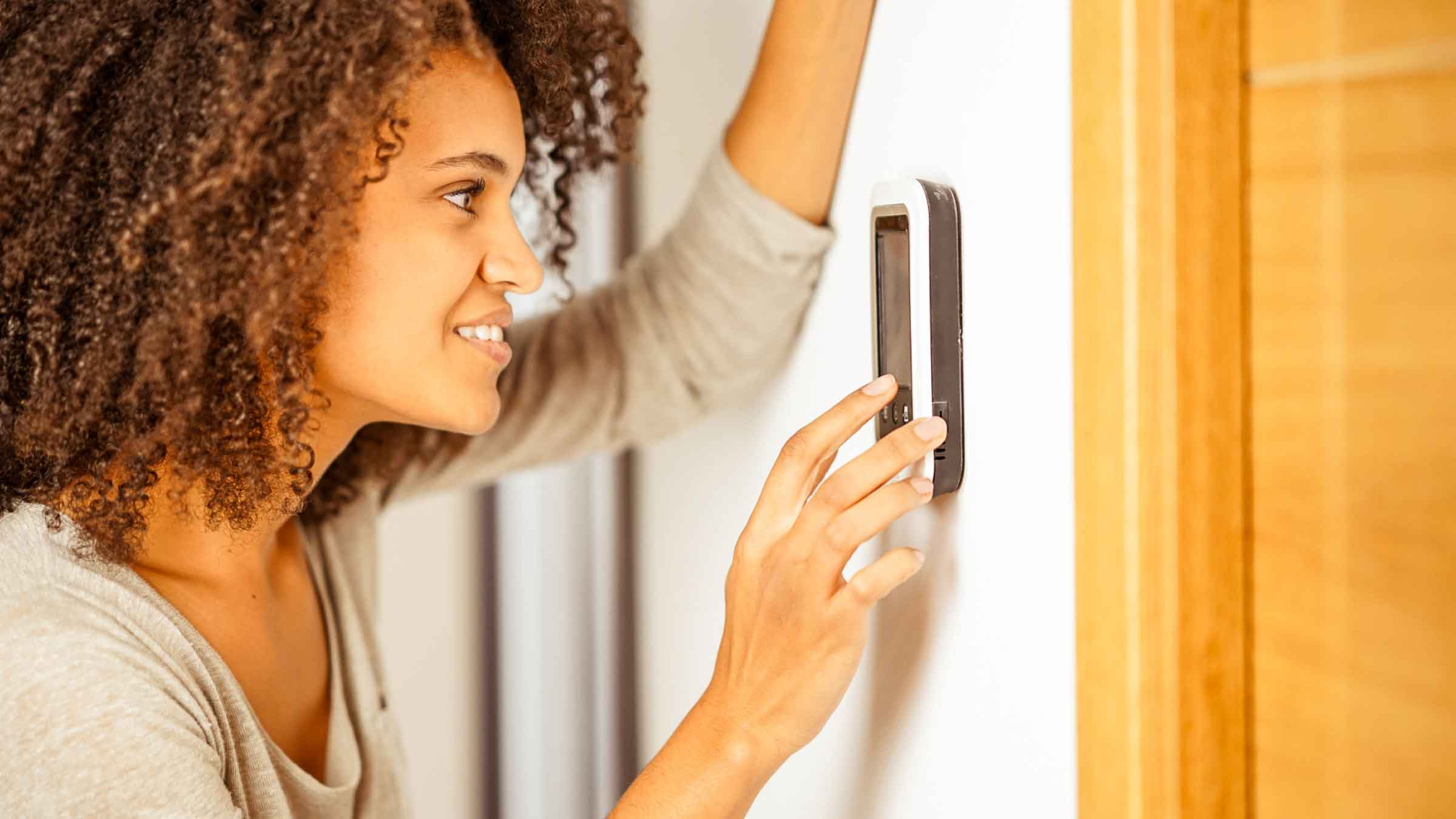 Woman leans against wall with her left arm and adjusts a smart home device with her right hand.