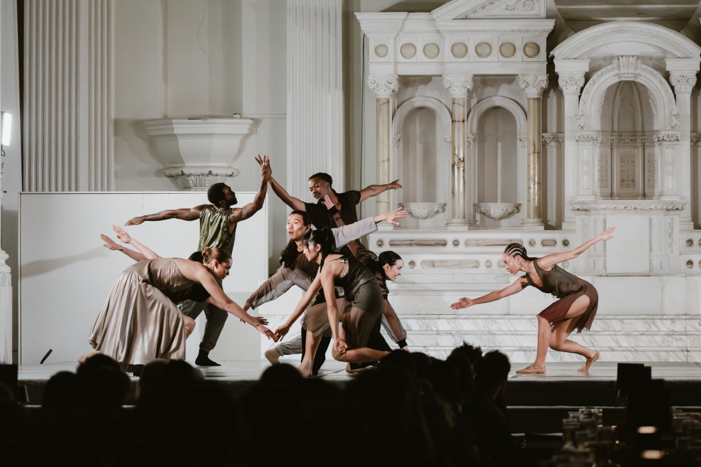 a group of dancers form a shape with their arms pointed in different directions, all dressed in neutral colors, at a performance at Vibiana in Downtown Los Angeles