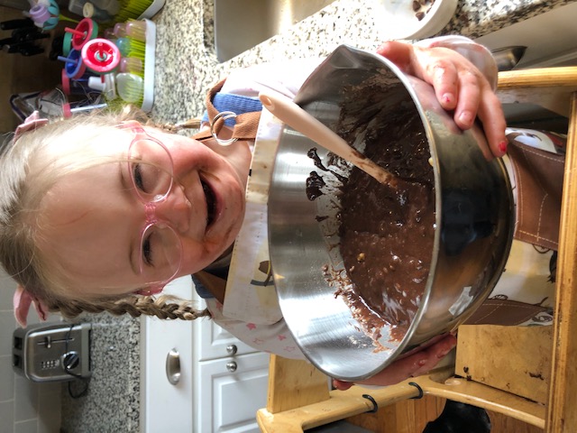 A little girl with Down syndrome wearing pink glasses smiling as she presents the bowl of chocolate cake mix that she just prepared