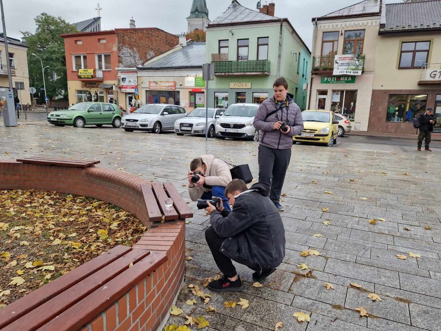 Fotografia jeszcze raz | Trzech chłopców z aparatami na kamiennej płycie Rynku. Dwóch chłopców kuca robiąc zdjęcie leżącej na deskach ławki szklanej kuli. W tle samochody i kamiemicznki..jpg