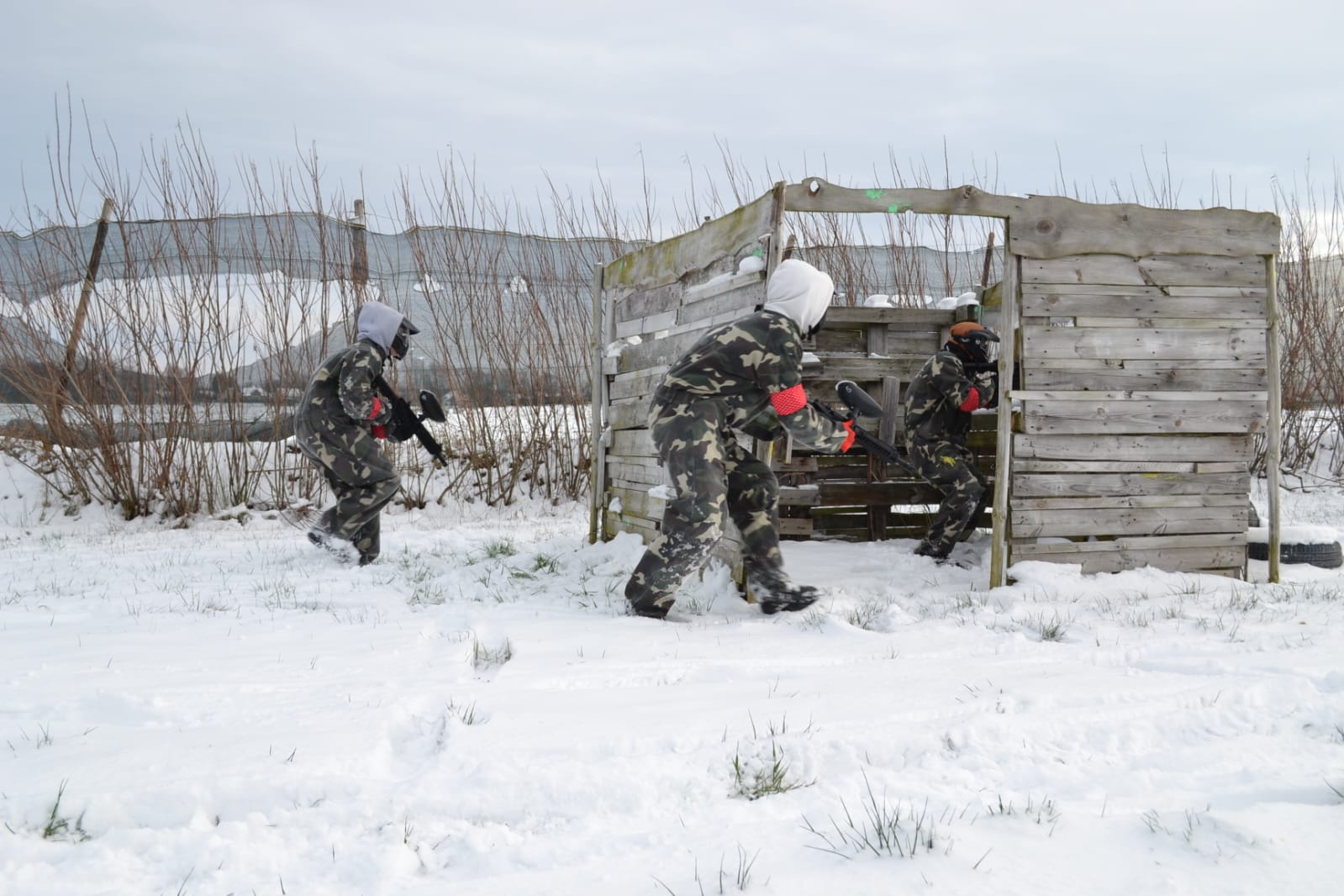 Wycieczka integracyjna klas Branżowej Szkoły I Stopnia w MOW w Lubaczowie | Trzy osoby w pełnym stroju do paintballa biegną pochylone z karabinkami w stronę ścianki z desek. Za nimi krzaki, dokoła świerzy śnieg.jpg